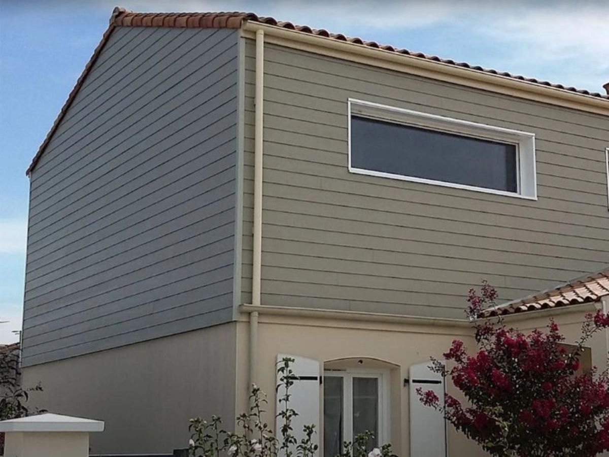 Étage en bardage bicolore vert et gris-bleu, rez-de-chaussée enduit beige, tuiles rouges.