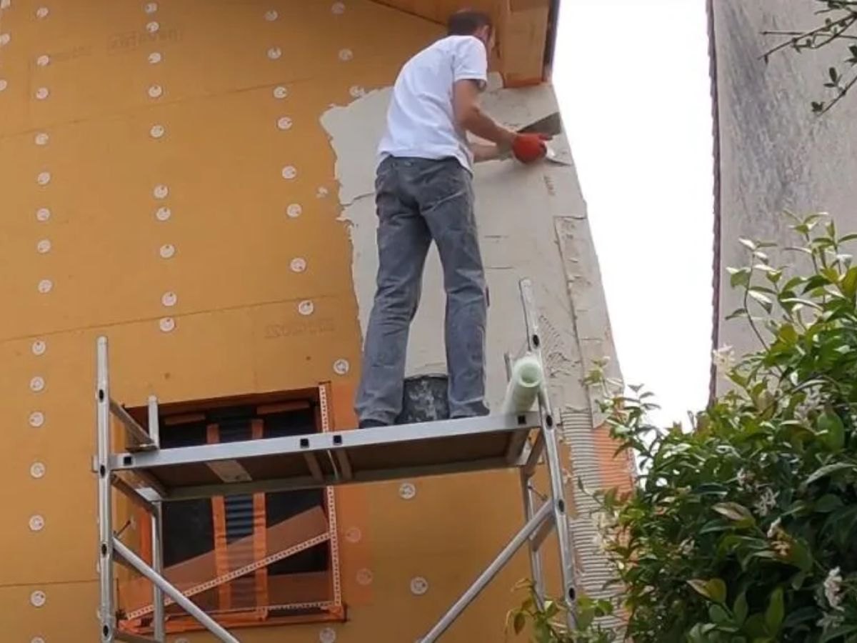 Ouvrier de profil sur échafaudage, talochant un enduit gris sur isolant orange.