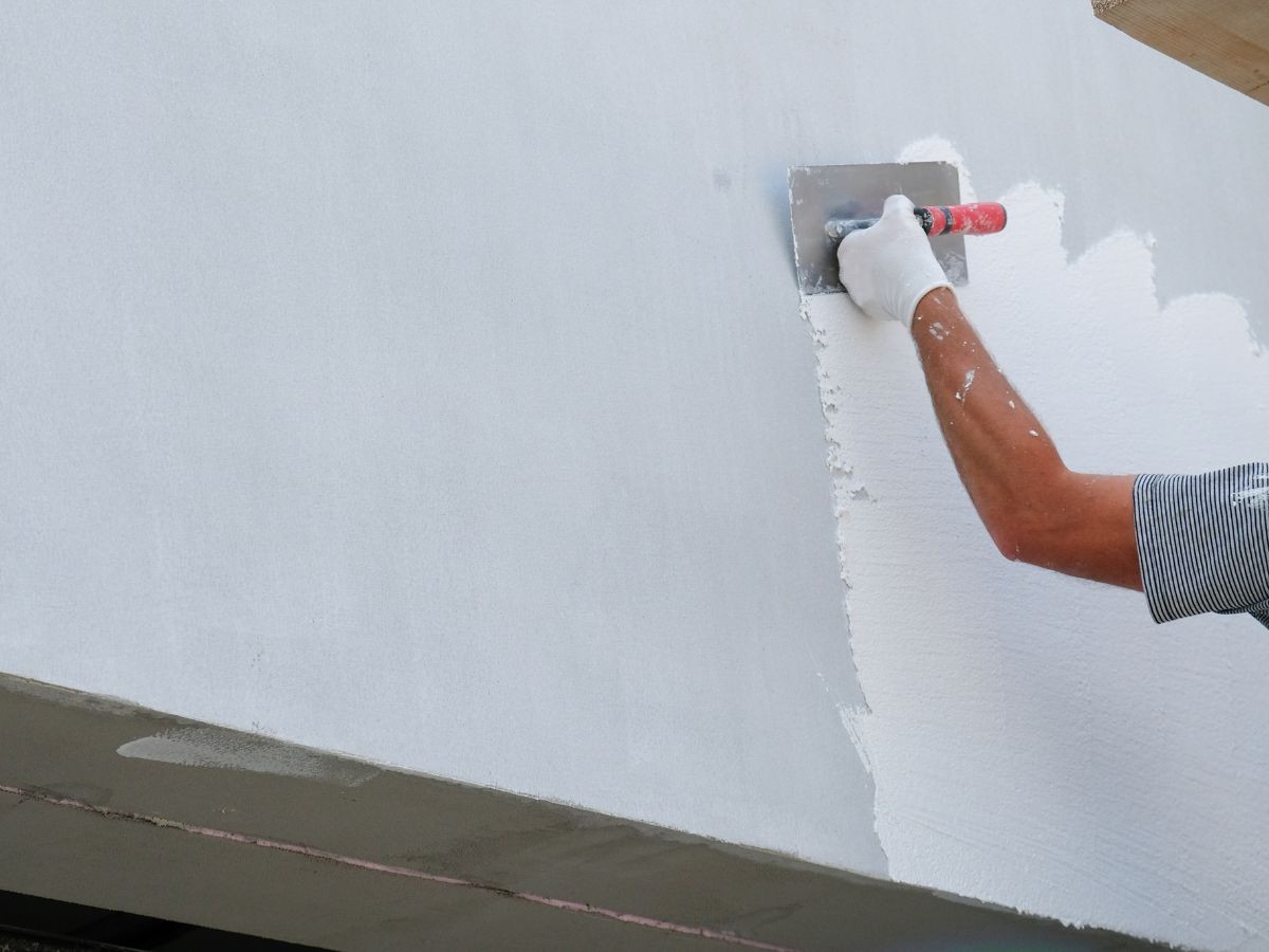 Bras ganté étalant du crépi blanc texturé sur sous-couche grise à la lisseuse, bord béton visible.