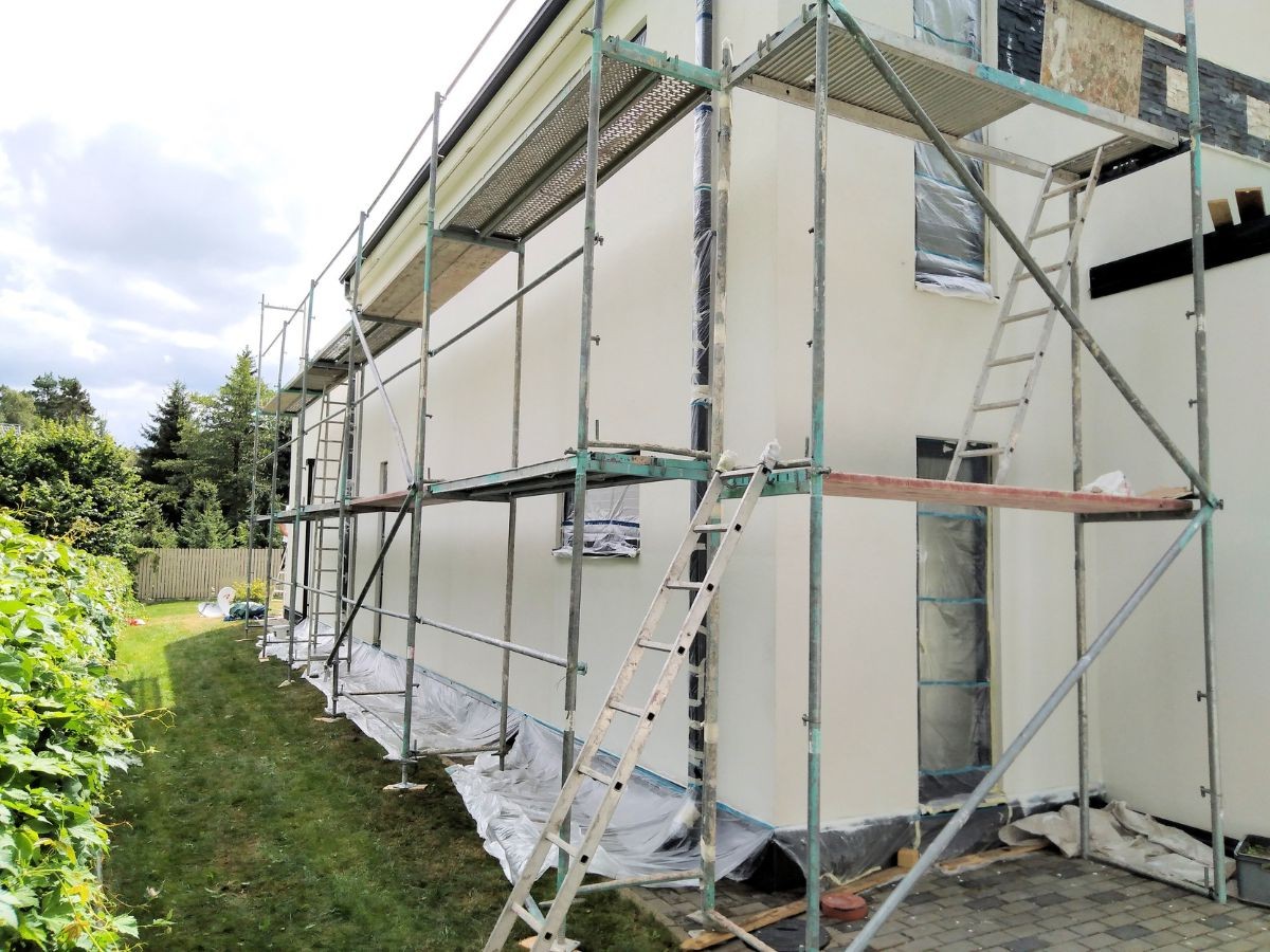 Long échafaudage le long d'une maison enduite crème, fenêtres bâchées, bâches blanches sur pelouse et allée.