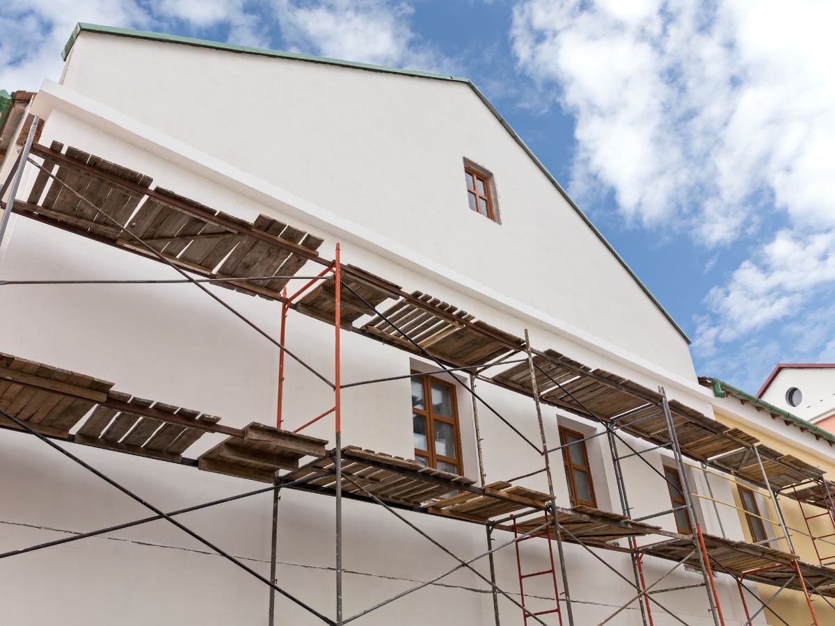 Échafaudage multi-niveaux avec planches bois devant pignon enduit blanc immaculé, fenêtres brunes sous ciel bleu.