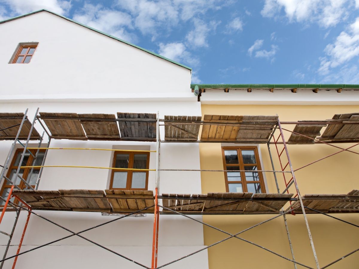 Chantier avec échafaudage métallique : moitié façade blanche, moitié jaune ochre, fenêtres bois et gouttière verte.