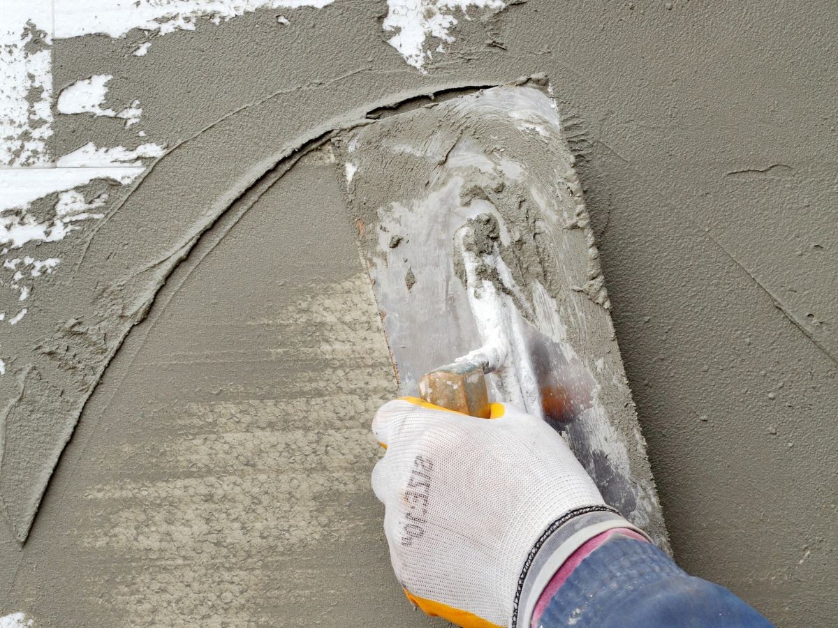 Gant orange talochant un enduit gris granuleux au couteau sur mur blanc écaillé, spirale fraîche visible.