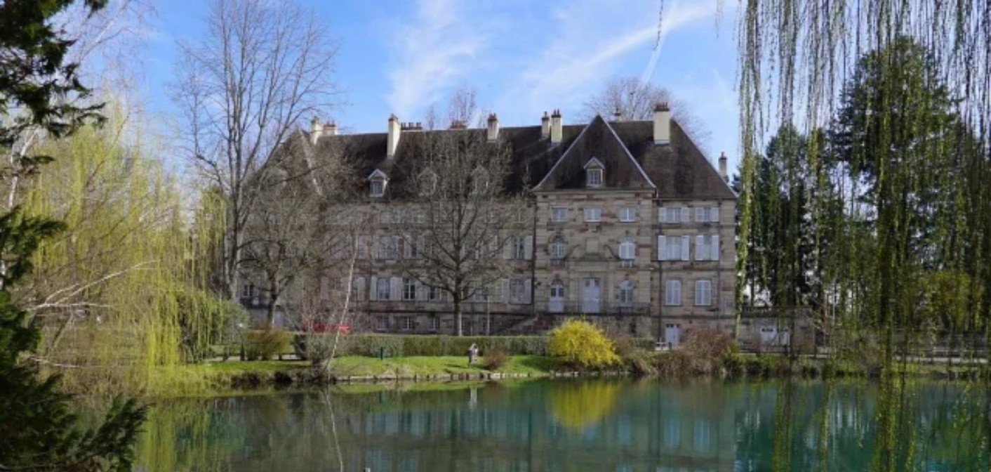 Manoir en pierre claire reflété dans un étang, saules et arbuste en fleurs.